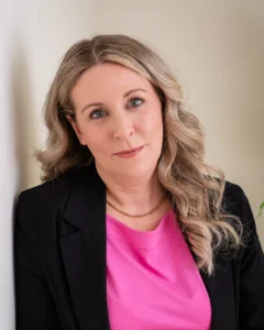Portrait of a professional woman with long, wavy blonde hair, wearing a black blazer over a bright pink top. She is leaning against a light-colored wall, looking confidently at the camera.
